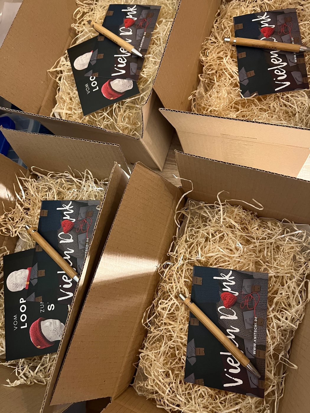 Several prepared KNITSCHI shipping boxes with wood wool, product cards, and ballpoint pens